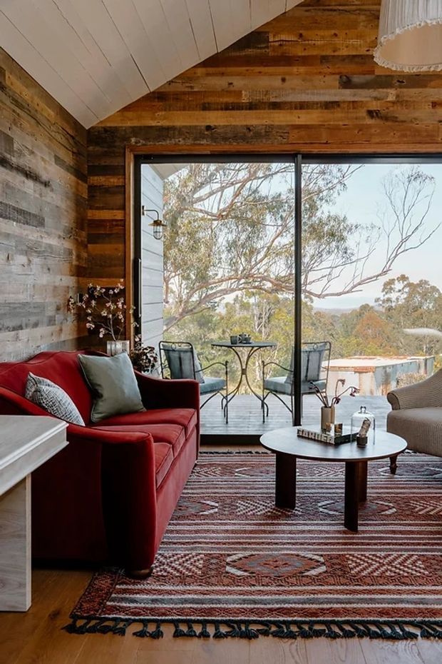 A warm, rustic living room with a red upholstered sofa, patterned area rug, round coffee table, and woven armchair. Reclaimed timber cladding covers the walls, while a white painted vaulted ceiling creates a distinctive angled roof form. Full-height black-framed glazing opens to a small outdoor deck with chairs and expansive views of surrounding trees and hills.