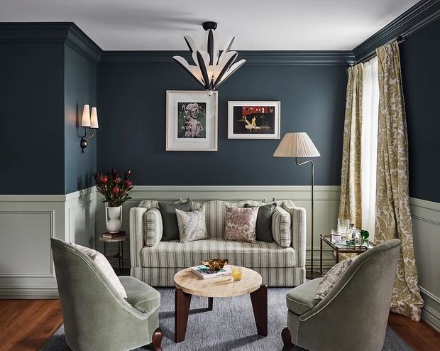 A refined sitting room with deep navy walls, white-painted wainscoting, decorative wall art, a striped upholstered sofa, two curved armchairs, and a round wood-and-stone coffee table. Patterned full-length curtains, layered lamps, timber flooring, and a sculptural pendant create a polished, traditional interior with a moody yet inviting atmosphere.