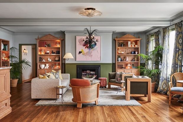 A richly layered, symmetrical living room with warm timber flooring, built-in wood shelving, a central fireplace, upholstered seating, tropical plants, patterned full-height curtains, and decorative wall moulding. The palette combines olive green, pale grey, terracotta, blush, and natural wood, creating an eclectic yet refined traditional interior.