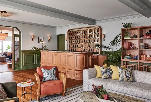 A richly detailed traditional living room and home bar area with warm timber flooring, green wainscoted walls, ornate ceiling mouldings, and an arched glazed doorway to an adjoining room. A built-in timber bar with an arched bottle display and shelving dominates the rear, while a boucle-style sofa, patterned cushions, armchair, coffee tables, plants, and layered rugs create a refined, inviting lounge.
