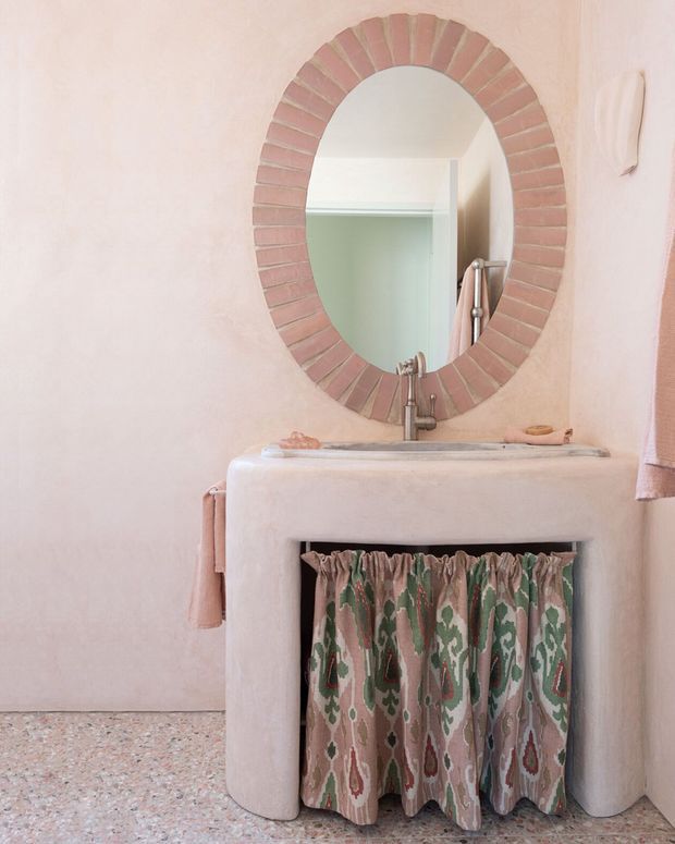 A compact powder-room vanity with a pale plaster or stucco finish, rounded integrated basin, and an oval mirror framed with radial pink-toned brick tiles. A patterned fabric curtain conceals the open vanity storage below, while a wall-mounted towel and terrazzo-style floor complete the softly coloured interior.