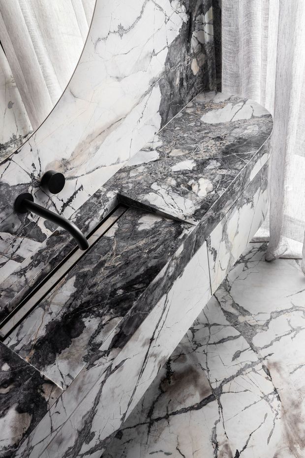 Close-up of a highly detailed marble bathroom vanity with an integrated basin and wall-mounted matte black tapware. The vanity, splashback, and floor are clad in dramatic white and grey veined natural stone, with sheer curtains beside a rounded mirror.
