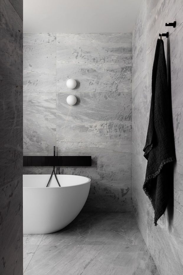 A compact, dramatic modern bathroom featuring a freestanding white bathtub, wall-mounted black tapware, black towel hooks and towel, and grey veined stone-look tiles covering the walls and floor. The space has a restrained monochrome palette and minimalist fittings.