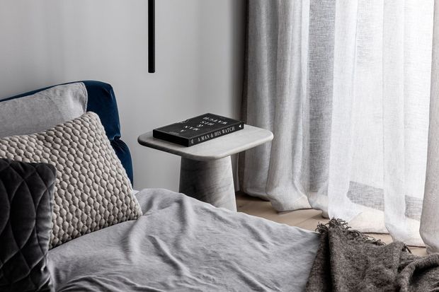 A softly lit bedroom corner with a grey upholstered bed, layered textured cushions and bedding, a small sculptural side table holding books, sheer floor-length curtains, and a dark vertical wall light. The restrained neutral palette and tactile fabrics create a calm, refined atmosphere.