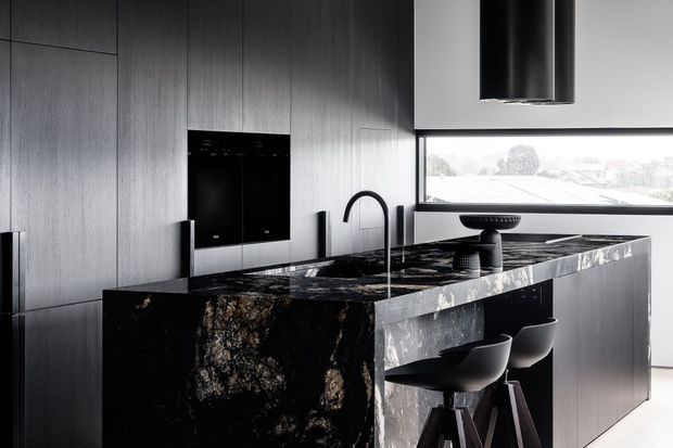 A sophisticated contemporary kitchen dominated by a large dark stone island with an integrated sink and curved black tap. The island includes seating for two at sculptural black stools, while full-height dark timber cabinetry lines the wall behind it. Black appliances and a large cylindrical rangehood reinforce the monochromatic, high-contrast palette. A wide horizontal window provides daylight and views beyond.
