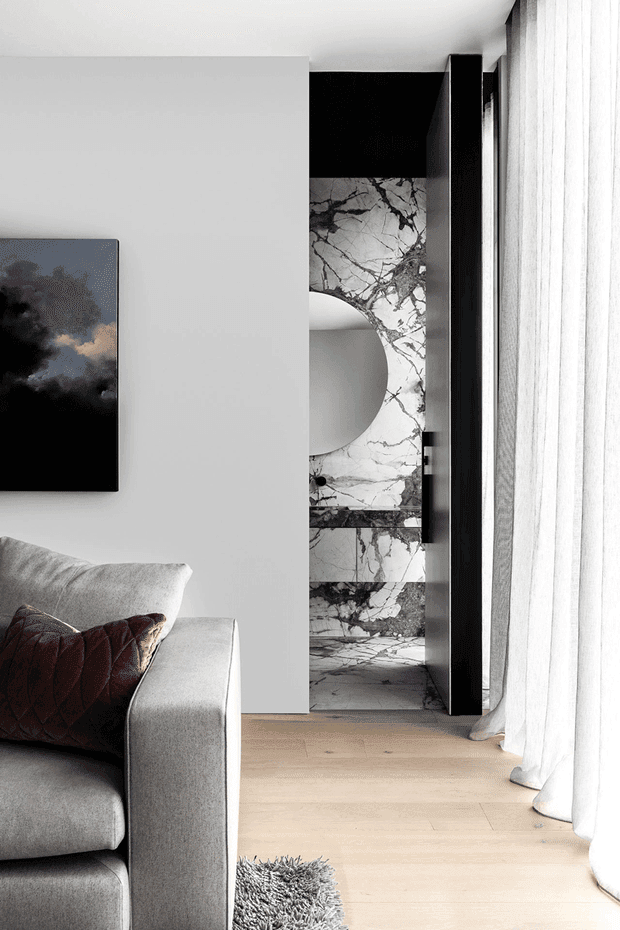 A refined contemporary living room with a light grey upholstered sofa, dark textured cushion, pale timber flooring, and sheer white curtains. A partially concealed marble-lined bathroom or vanity zone is visible behind a white partition, framed by black built-in joinery and a dramatic dark artwork.