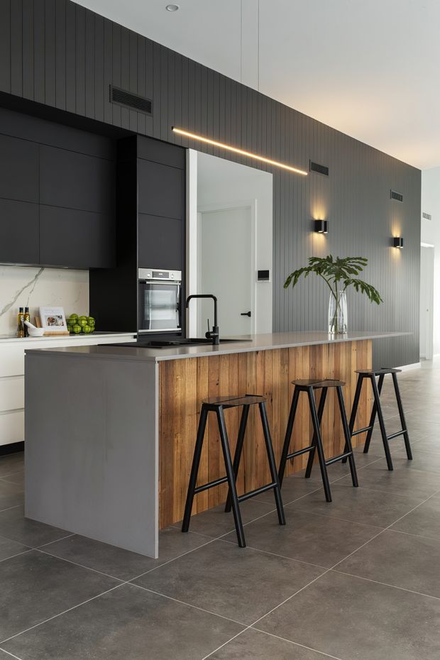Contemporary open-plan kitchen featuring a long island with a dark grey side panel, warm timber-panelled seating face, integrated sink and black tap, and four black bar stools. Matte dark cabinetry, white marble-look splashback, large-format grey floor tiles, and a vertically grooved dark feature wall are complemented by linear and wall-mounted lighting.