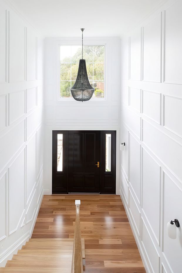 Bright, tall entry stair hall viewed from above, featuring white decorative wall panelling, warm timber flooring and staircase, a dark timber front door with narrow glazed sidelights, a large upper window, and a striking black pendant light.