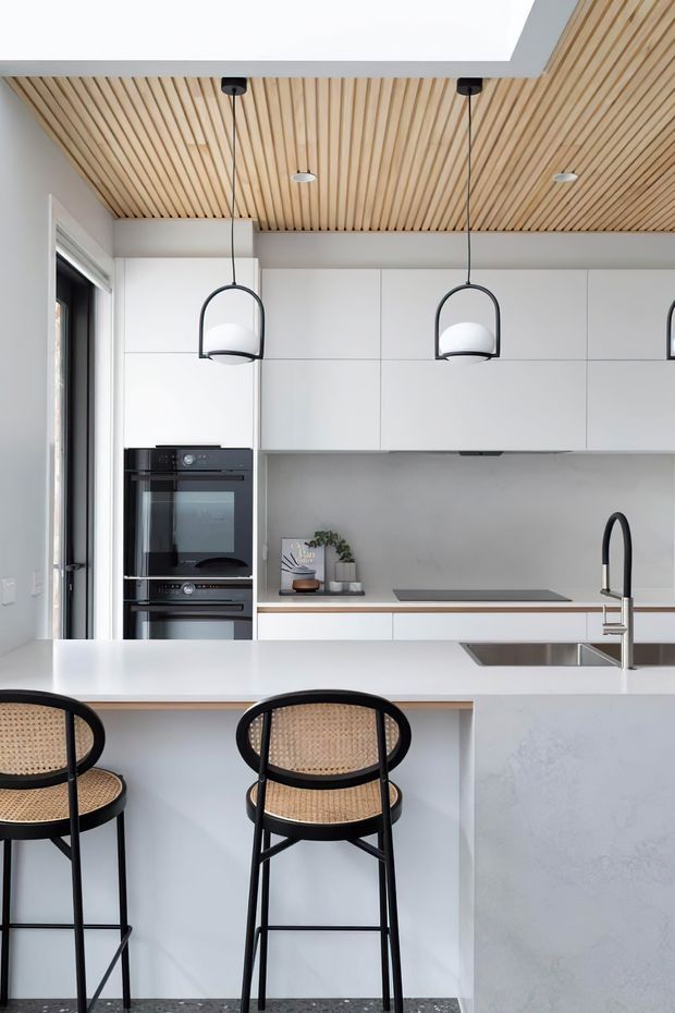 Bright contemporary kitchen featuring a large white island with integrated sink and black gooseneck tap, white flat-panel cabinetry, a grey backsplash, built-in black ovens, and a timber slatted ceiling. Black-framed pendant lights and black rattan bar stools provide contrast, while a dark terrazzo-like floor completes the space.