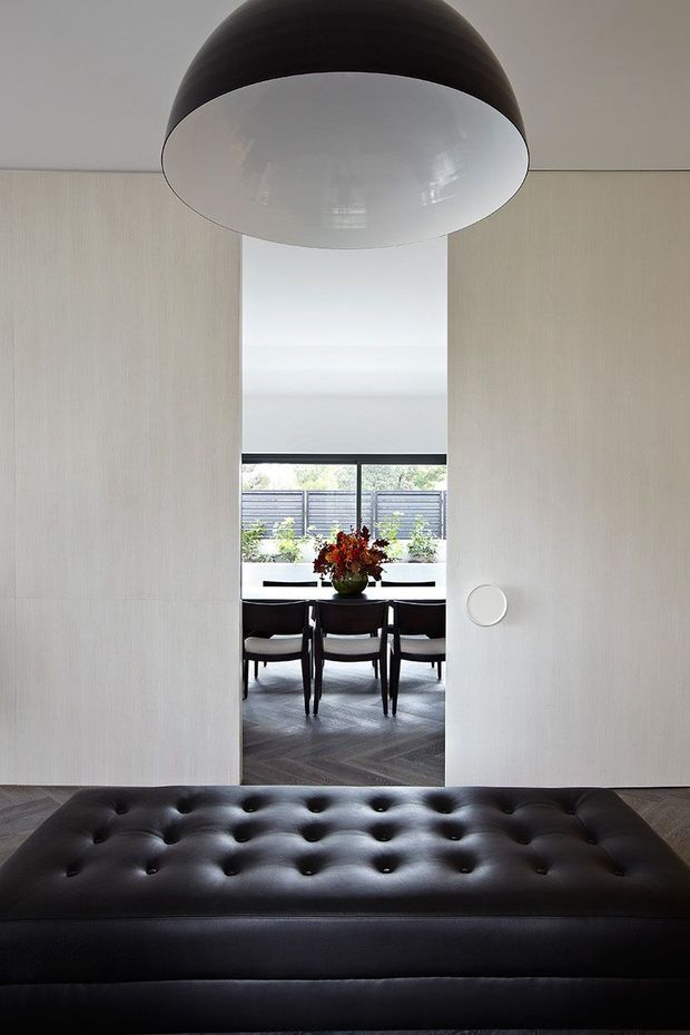 A refined interior view through a wide doorway, featuring a black tufted leather bench in the foreground, pale textured wall panels, and a large black-and-white pendant light. Beyond the opening is a dining area with a dark table and chairs, abundant natural light, and patterned dark flooring. The composition is minimalist and monochromatic with strong geometric forms.
