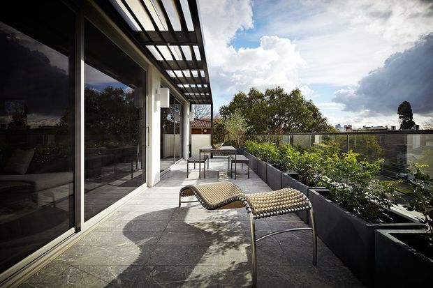 A spacious rooftop or upper-level terrace adjoining a modern interior through full-height glazing. The terrace has dark stone or concrete paving, a slatted pergola or sunshade overhead, built-in raised planters, outdoor dining furniture, and sculptural lounge chairs. Glass balustrades provide open views toward surrounding trees and neighbouring buildings under a partly cloudy sky.