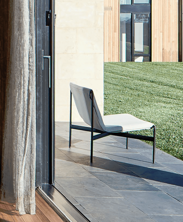 A close architectural view from inside looking through full-height glazing toward a minimalist outdoor terrace. A sculptural black-framed lounge chair sits on large grey stone paving beside a neatly clipped green hedge. The building exterior combines pale rendered or stone-like wall surfaces, vertical light timber cladding, and dark aluminium window and door frames. A sheer curtain and timber interior floor are visible in the foreground.