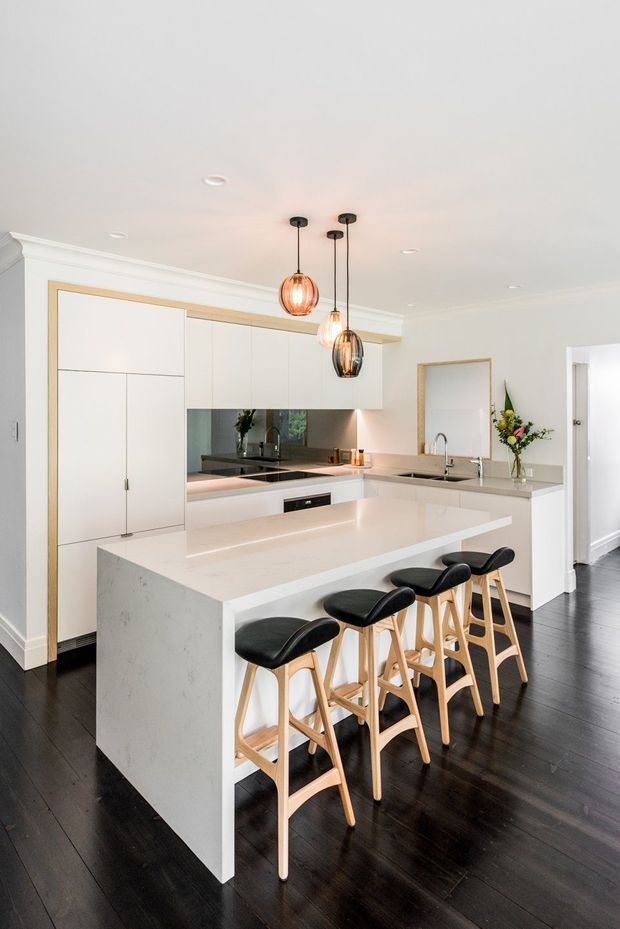 Bright contemporary kitchen with a large white waterfall island, integrated white cabinetry, pale stone benchtops, mirrored splashback, dark timber flooring, and four timber bar stools with black seats. Three pendant lights hang above the island, while a sink and additional preparation area sit along the rear benchtop. A doorway and floral arrangement are visible to the side.