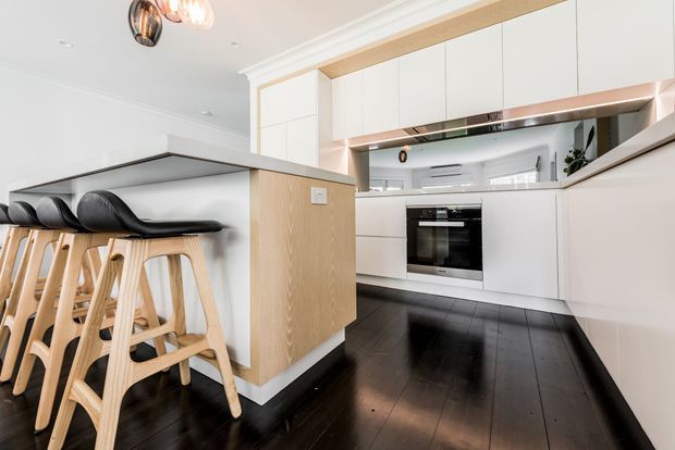 Bright contemporary kitchen viewed from the adjoining living area, featuring a white U-shaped kitchen with integrated cabinetry, a black oven, reflective splashback, pale timber island panel, dark timber flooring, and a breakfast bar with sculptural timber stools. Recessed lighting and a decorative pendant add a refined modern feel.