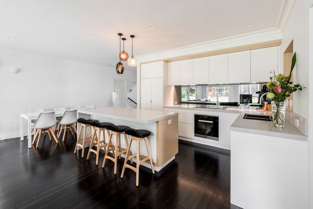 A bright, spacious contemporary kitchen and dining area with a large white island, integrated cabinetry, dark timber flooring, and seating for several people. White dining chairs and black-and-timber bar stools surround the island, while pendant lights hang above. A mirrored splashback reflects the kitchen and surrounding windows, and subtle ceiling cornices add a refined architectural detail.