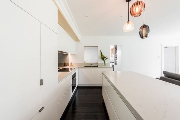 Bright contemporary kitchen viewed from the main work area, featuring extensive white handleless cabinetry, a large pale stone island benchtop, dark timber flooring, integrated appliances, and three contrasting pendant lights. A small adjoining living area is visible at right.