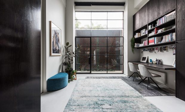 A refined home office in a narrow, high-ceilinged space with a large black-framed glazed wall, built-in dark timber shelving and desk, two grey upholstered chairs, a pale patterned rug, indoor plants, framed artwork, and a rounded blue ottoman. The room combines strong architectural glazing with dark joinery and a light neutral palette.