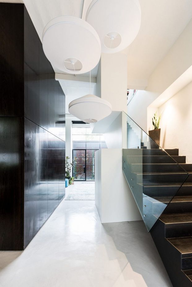 Bright contemporary entry hall with a dramatic open staircase. The space features dark full-height built-in cabinetry or wall panelling, a polished light concrete floor, white walls, glass stair balustrades, and multiple large round pendant lights. A skylight and glazed rear doorway bring in natural light, while indoor plants add a small organic accent.
