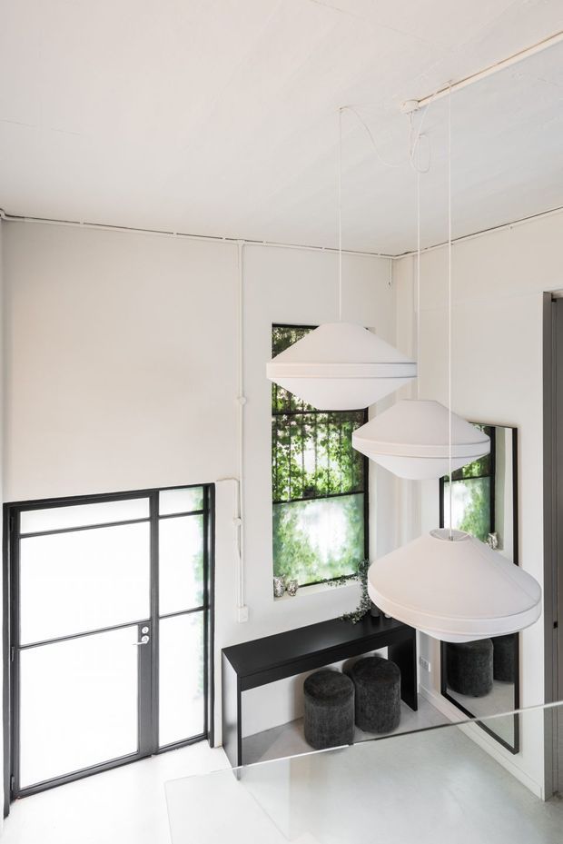 Bright minimalist interior entryway with white walls and ceiling, exposed surface-mounted electrical conduits, black-framed glazed door and windows, a slim black console table, round dark upholstered stools, and several sculptural white pendant lights. A large mirror reflects the seating and pendant fixtures, while greenery is visible through the windows.