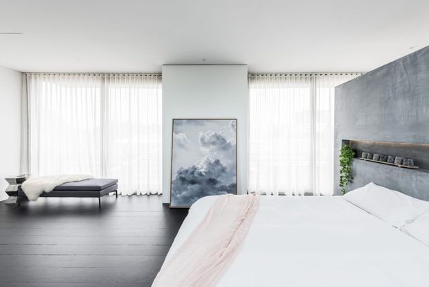 Bright contemporary bedroom with a low-profile bed, white bedding, pale pink throw, dark timber flooring, sheer floor-to-ceiling curtains, and a textured grey feature headboard incorporating a narrow shelf and greenery. A framed artwork leans against the central wall, while a chaise or bench sits near the windows.