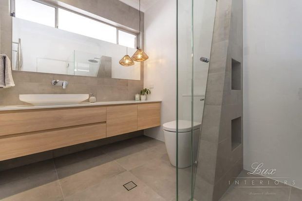 Contemporary bathroom with a long floating timber vanity, white vessel basin, wall-mounted fixtures, large warm grey wall and floor tiles, frameless glass shower enclosure, concealed shower niches, and a freestanding toilet. A large mirror and high horizontal window bring brightness into the space, while pendant lights add a warm decorative accent.
