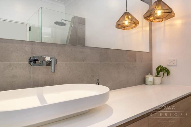 Contemporary bathroom featuring a freestanding white oval bathtub, wall-mounted chrome tapware, large-format grey wall tiles, a long pale benchtop with integrated basin area, frameless glass shower screen, and warm geometric pendant lights.