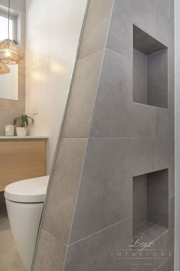 Contemporary bathroom featuring a sculptural angled partition clad in large-format grey tiles, with recessed niches, a wall-mounted timber vanity, white toilet, and geometric pendant lighting.