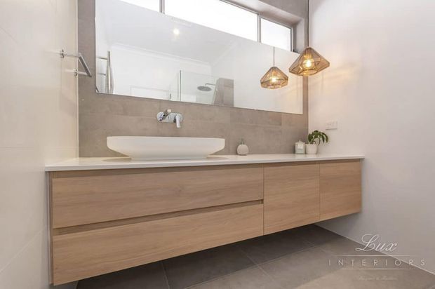 Contemporary bathroom vanity with a long floating timber-look cabinet, white countertop and vessel basin, large mirror, grey tiled splashback, and warm pendant lighting. The bright, clean space has a refined neutral palette and integrated storage.