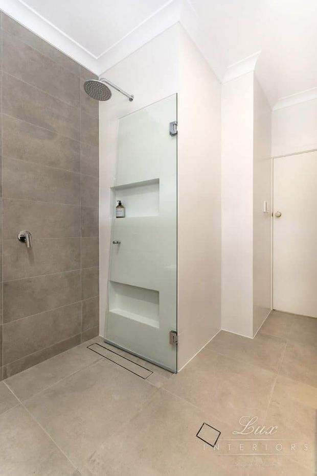 Bright, contemporary bathroom featuring a walk-in shower with large grey-beige wall and floor tiles, a frameless glass shower screen with built-in recessed niches, a rainfall showerhead, white painted walls with decorative cornices, and a simple white door.