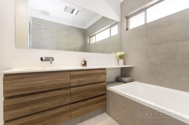 Contemporary bathroom with a double timber-look vanity, white countertop and integrated basin, large frameless mirror, pale grey tiled walls, and a built-in bathtub. High horizontal windows bring in natural light, creating a clean, bright and neutral space.