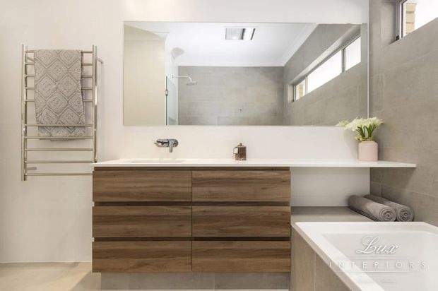 Bright contemporary bathroom featuring a floating double-width timber vanity with integrated white benchtop and basin, a large wall mirror, wall-mounted chrome tapware, grey tiled walls and floor, a freestanding bathtub, heated towel rail, and a narrow high window. The room has a clean, refined, neutral aesthetic with warm wood cabinetry.