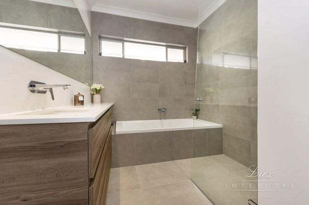 Contemporary bathroom featuring a wall-mounted timber-look vanity with white benchtop, frameless glass shower enclosure, integrated bathtub, and large-format grey-beige wall and floor tiles. High horizontal windows provide natural light, with clean white ceiling trim and minimal chrome tapware.