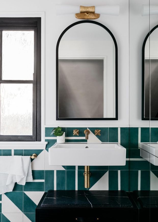 A compact, highly styled bathroom featuring a wall-mounted white rectangular basin, black-framed arched mirror, black-framed window, brass wall-mounted tapware and lighting, and striking teal geometric tiled walls. A black vanity with a dark stone top sits below the basin, while the mirror reflects additional cabinetry or wall surfaces.