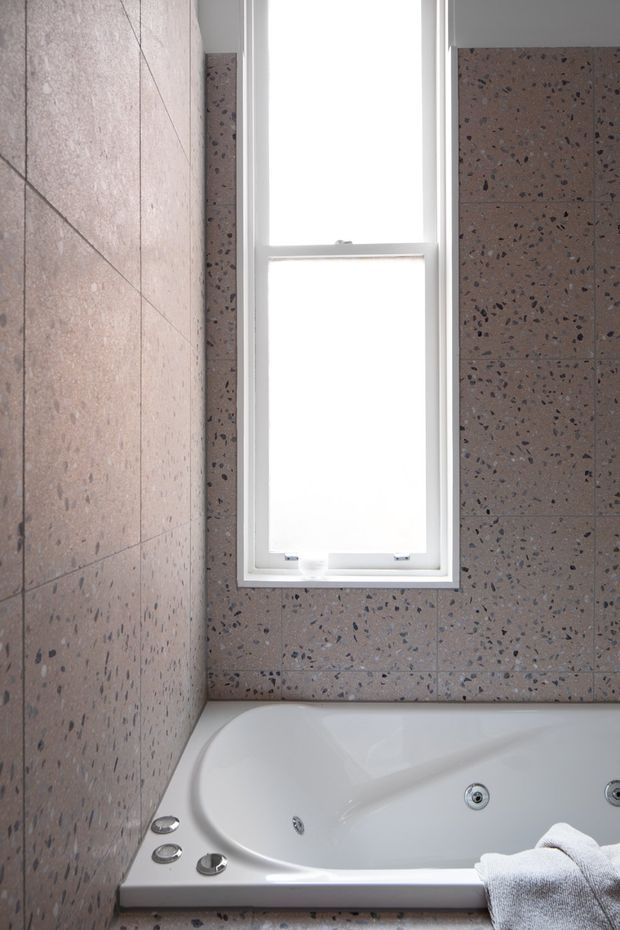 Close-up view of a bathroom featuring a white corner whirlpool bathtub beneath a tall frosted or overexposed window. The walls are clad in large pinkish-grey terrazzo tiles with dark aggregate, and the window has a white painted frame. Chrome bath jets and controls are visible, with a folded white towel resting on the tub edge.