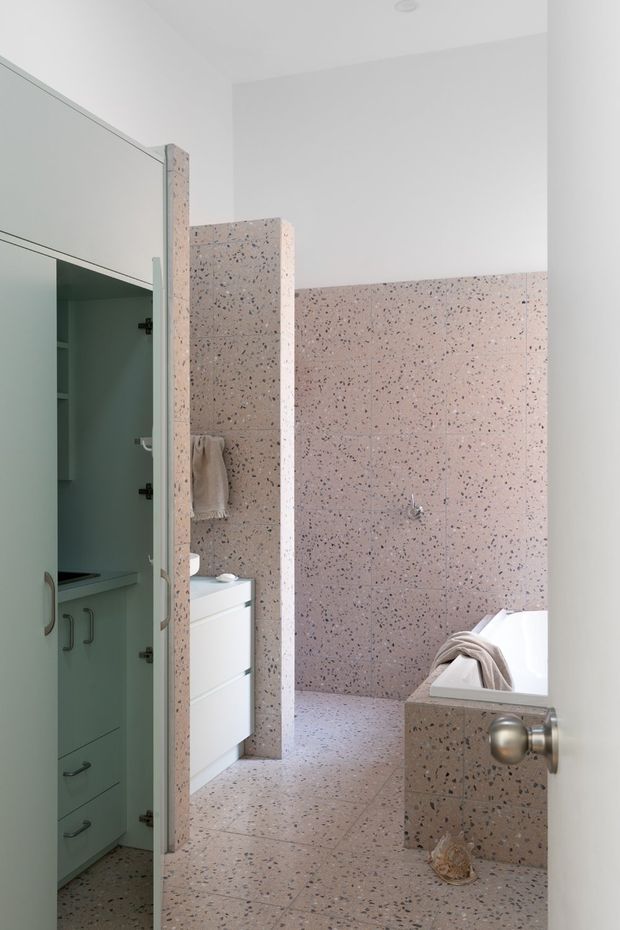 Bright contemporary bathroom viewed through an open doorway, featuring extensive pink terrazzo wall and floor tiling, a freestanding or partially enclosed bathtub, white vanity cabinetry, and a muted sage-green built-in storage area visible to the left. A terrazzo partition separates the vanity from the bath, creating a clean, geometric layout.