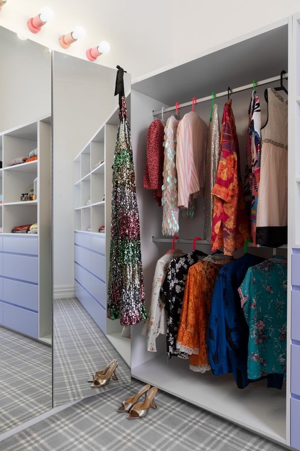 A bright, custom walk-in wardrobe filled with hanging clothing, open shelving, and pale lavender drawer cabinetry. Full-height mirrored wardrobe panels reflect the storage area, while a grey checked carpet and vanity-style globe lights create a dressing-room feel.