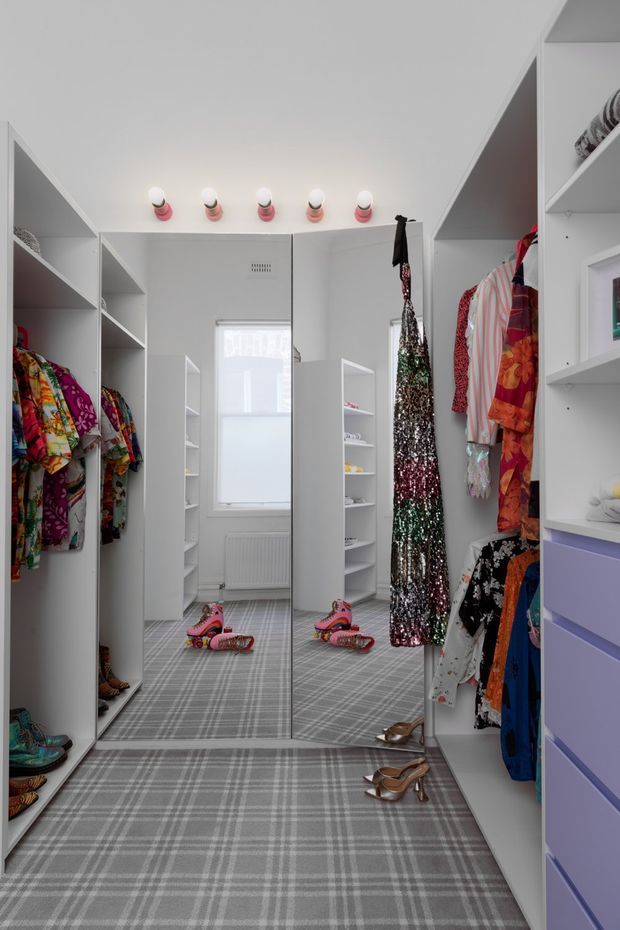 A bright, highly organised walk-in wardrobe or dressing room with extensive white open shelving and hanging rails on both sides. Colourful clothing, shoes and accessories are displayed throughout, with full-height mirrored doors at the rear reflecting the room. The room has a grey plaid carpet, white cabinetry, a small window, and decorative wall-mounted lights above the mirror.