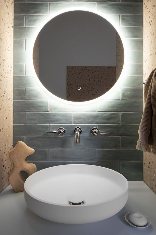 A compact, contemporary bathroom vanity with a white round vessel basin set on a grey countertop. The wall behind is clad in blue-grey rectangular tiles, framed by terrazzo-like speckled wall surfaces. A large round backlit mirror dominates the composition, with exposed chrome wall-mounted tapware beneath it. A small soap dish and sculptural wooden object sit on the vanity.