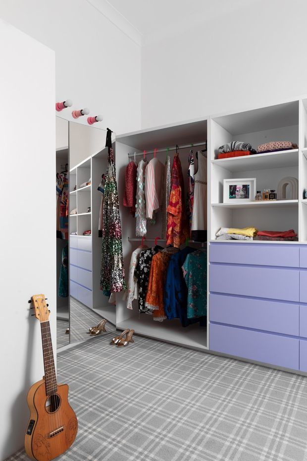 Bright, built-in walk-in wardrobe with open hanging rails, shelving, and lavender drawer cabinetry. The room has white walls and joinery, grey plaid carpet, full-height mirrors, and colourful clothing and accessories. A guitar and shoes are visible in the foreground, adding a playful residential character.