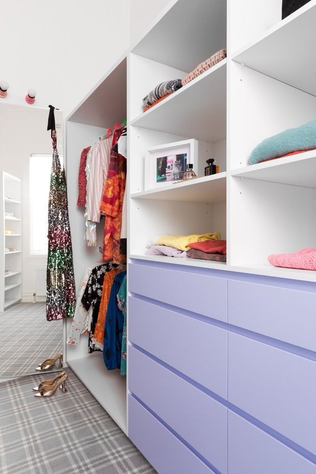 Bright custom walk-in wardrobe with white open shelving, hanging rails filled with colourful clothing, lavender built-in drawers, folded garments, accessories, and a grey plaid carpet. The space is compact but highly organised, with a playful fashion-focused palette.