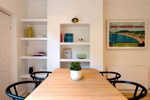Bright, warm dining area with a light timber rectangular dining table and black spindle-back chairs. Built-in white shelving and recessed display niches line the walls, holding books and decorative objects, while a framed vintage travel poster and white radiator add character.