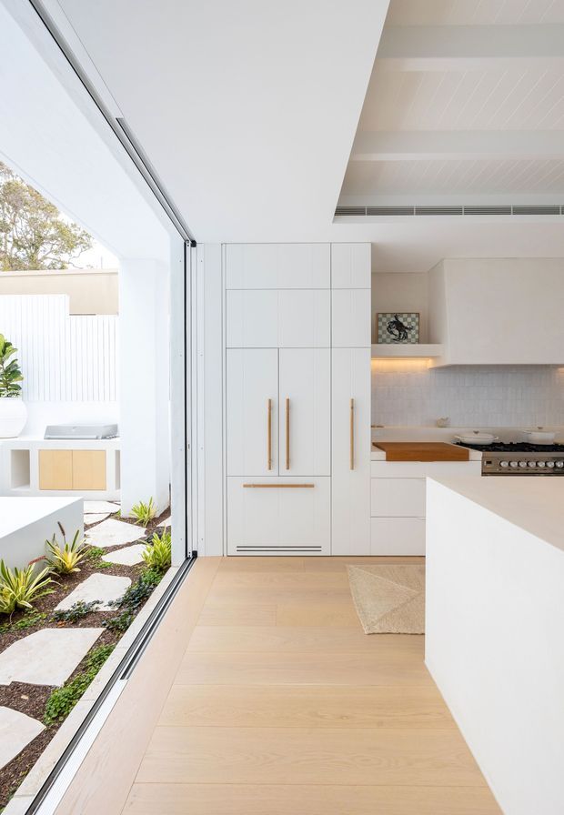 Bright contemporary kitchen with a minimalist white finish, integrated full-height cabinetry with slim brass-toned handles, a pale timber floor, white island, tiled splashback, and a timber benchtop section. Full-height sliding glass doors open directly to a small landscaped courtyard with stepping stones and built-in outdoor cabinetry. The ceiling features white painted beams and linear air-conditioning grilles.