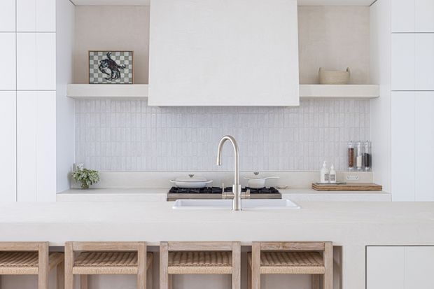 Bright, minimalist kitchen with a large white island, integrated gas cooktop, tall curved tap and undermount sink. Full-height white cabinetry frames open display niches, while a textured white tiled splashback sits beneath a concealed rangehood. Light timber woven-seat stools line the island, with restrained neutral styling and small decorative objects.
