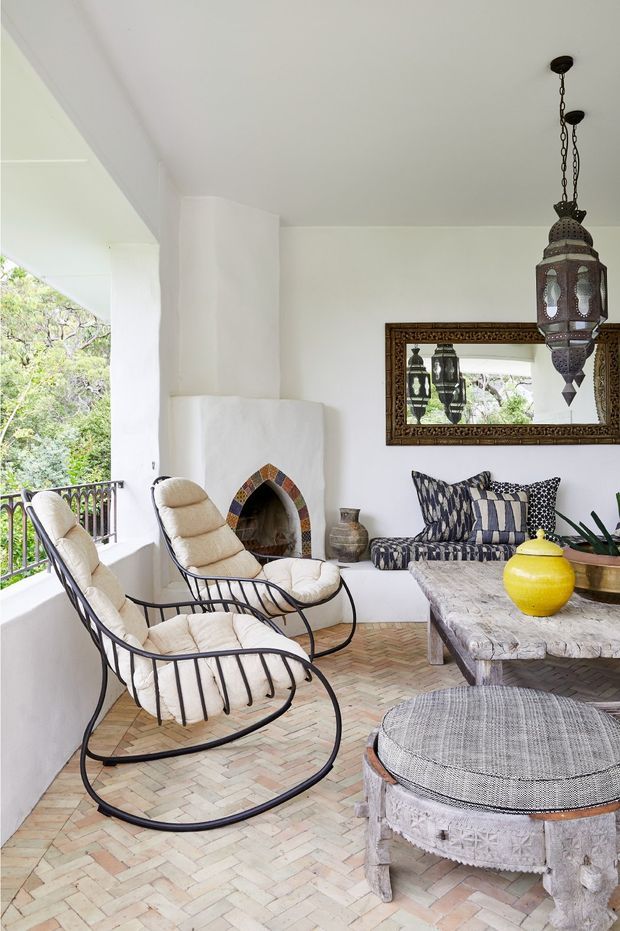 An airy covered outdoor lounge or enclosed terrace with white plaster walls, a small arched fireplace, black metal rocking lounge chairs, a patterned built-in sofa, a rustic stone coffee table, and a round upholstered ottoman. A decorative Moroccan-style pendant light and ornate wall mirror add character. The floor is laid in a pale herringbone brick pattern, with greenery visible beyond the balcony railing.