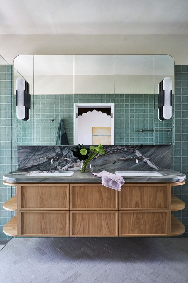 A refined double-vanity bathroom featuring a large mirrored wall cabinet, twin black-and-white wall sconces, emerald green square wall tiles, a dramatic dark veined stone benchtop and splashback, and a floating warm timber vanity with fluted rounded side shelves. The floor is finished in pale grey herringbone tile, and a decorative doorway and artwork are reflected in the mirror.