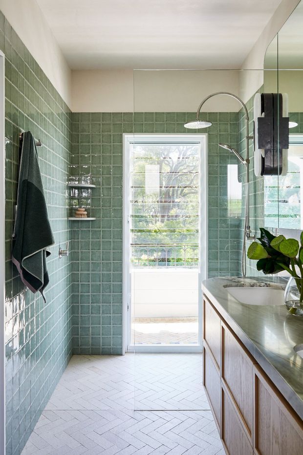 Bright, narrow contemporary bathroom with sage green glossy square wall tiles, white herringbone floor tiles, a timber vanity with a pale stone benchtop, integrated basin, large wall mirror, and a walk-in shower with curved chrome fittings. A louvred glazed door provides strong natural light and views to greenery outside.