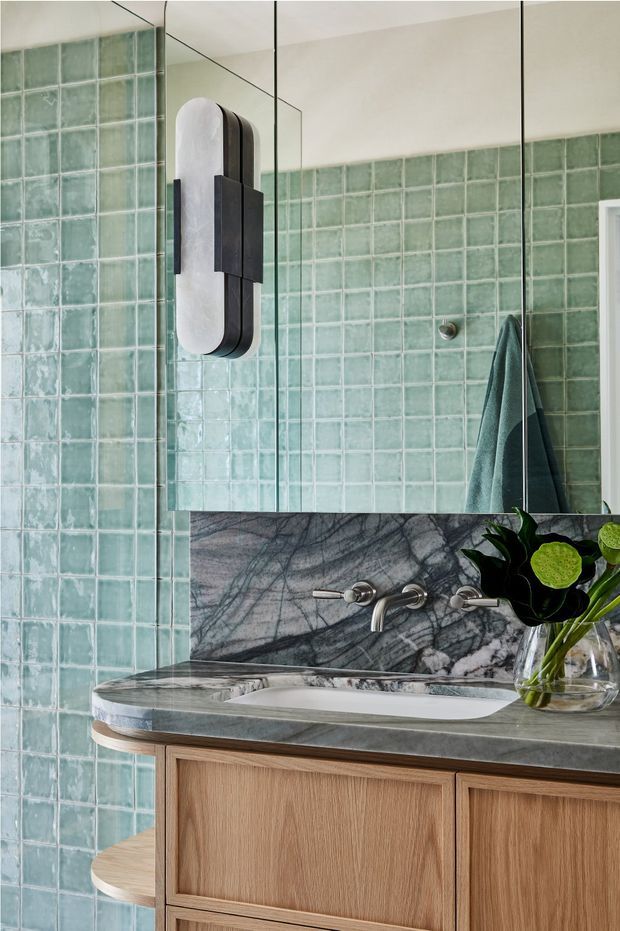 Close-up view of a refined bathroom vanity with a floating or wall-mounted brushed timber cabinet, grey veined natural stone benchtop and integrated basin. The wall and shower enclosure are lined with glossy, irregular pale green square tiles, complemented by a matching green towel. A large mirror, sculptural wall light, wall-mounted brushed metal tapware, and a small glass vase with greenery complete the setting.