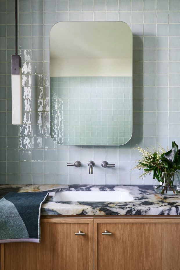 A stylish bathroom vanity with a freestanding bathtub set into a timber vanity cabinet. The walls are clad in pale blue-green square tiles, complemented by a large rounded-rectangle mirror, wall-mounted chrome tapware, and a striking veined marble benchtop. A folded dark teal towel and leafy floral arrangement add colour and texture.