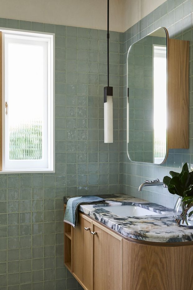 A compact, thoughtfully designed bathroom with sage-green square wall tiles, a warm timber vanity, and a striking natural stone countertop with an integrated white basin. A rounded-rectangle mirror, slender pendant light, chrome wall-mounted tap, and frosted window provide functional and decorative detail. The space feels bright, calm, and refined.