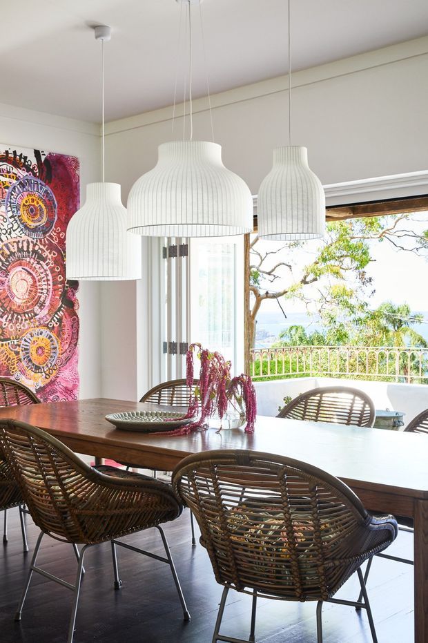 A bright dining room with a large dark timber dining table surrounded by woven rattan-style chairs. Four white ribbed pendant lights hang above the table, while colourful patterned artwork adds a bold accent on the wall. Large glazed doors open to a balcony or terrace with a leafy outdoor view. The room has dark timber flooring and a light neutral ceiling and walls.